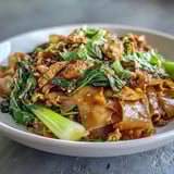 A close-up of Authentic Thai Pad See Ew reveals charred wide rice noodles, scrambled egg ribbons, and crisp gai lan in a fragrant stir-fry.  