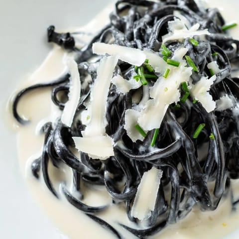 A close-up of Squid Ink Pasta twirled in a rich, glossy cream sauce, topped with Parmesan cheese and served on a white plate.
