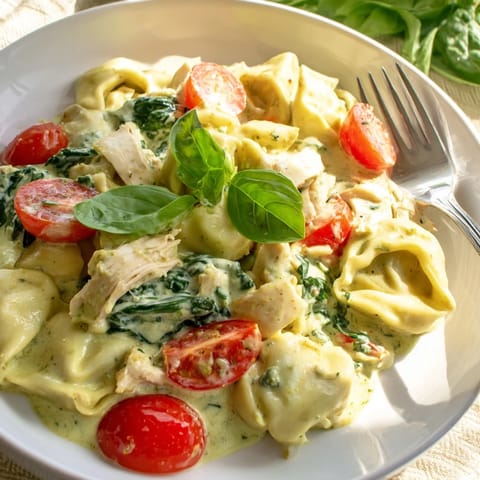 A close-up of cheesy tortellini coated in creamy pesto sauce, garnished with fresh basil and cherry tomatoes.