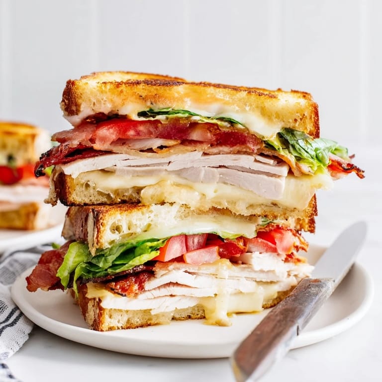 A close-up of the Turkey Club Grilled Cheese shows layers of melted Swiss cheese, savory turkey, crunchy bacon, and vibrant lettuce between crispy sourdough.  