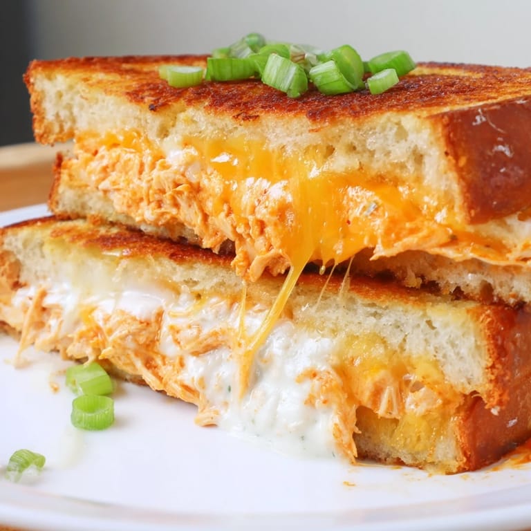 Buttery toasted bread holds melted cheese and tangy buffalo chicken, served with crisp green onions on a plate.